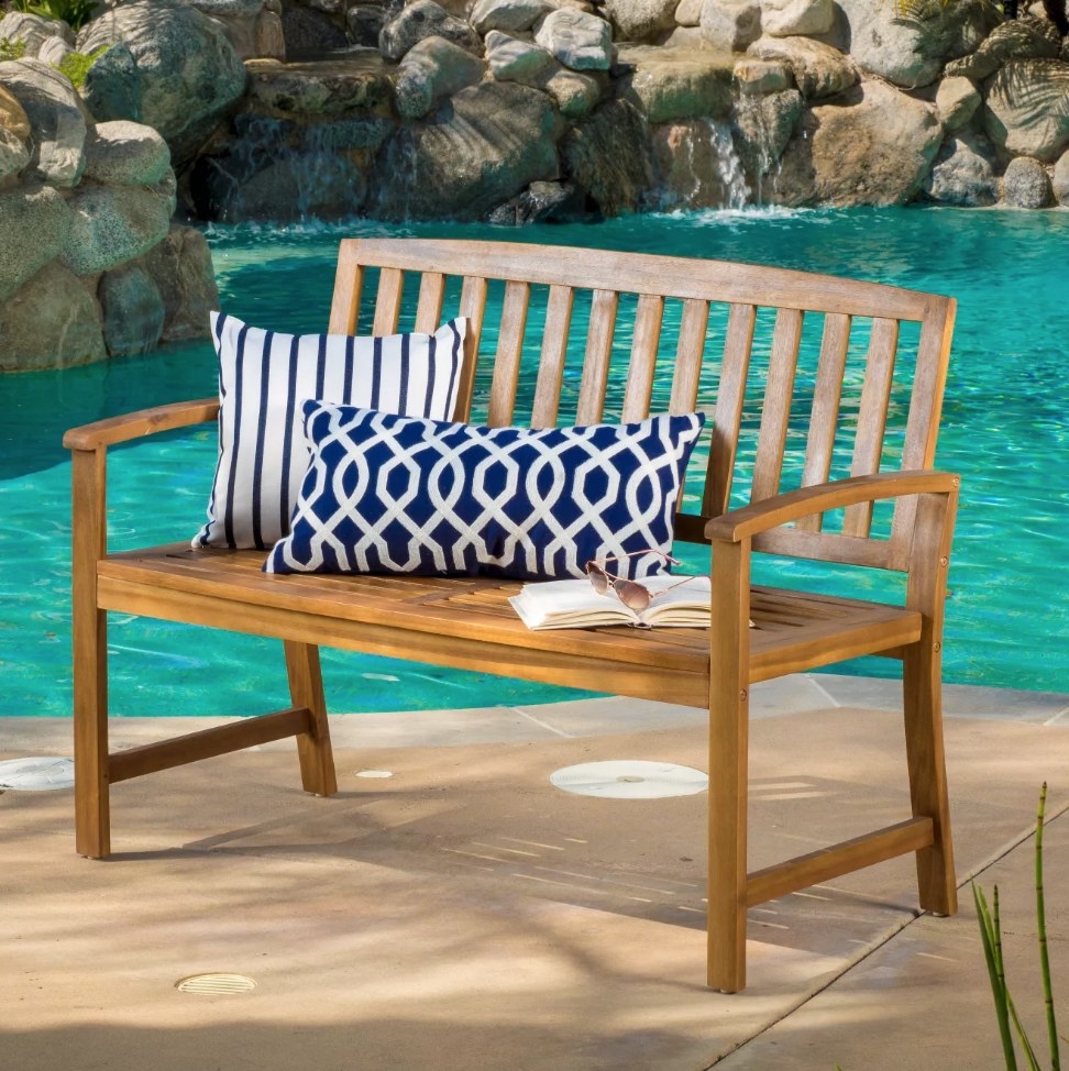 A brown, wooden bench outdoors by a pool with blue/white throw pillows atop