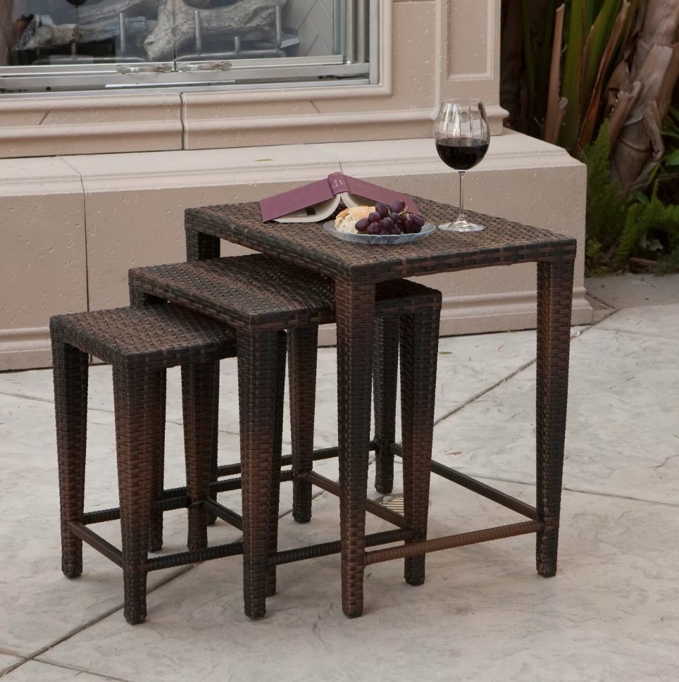 A set of 3 brown, wicker nesting tables on a patio with food and drinks atop