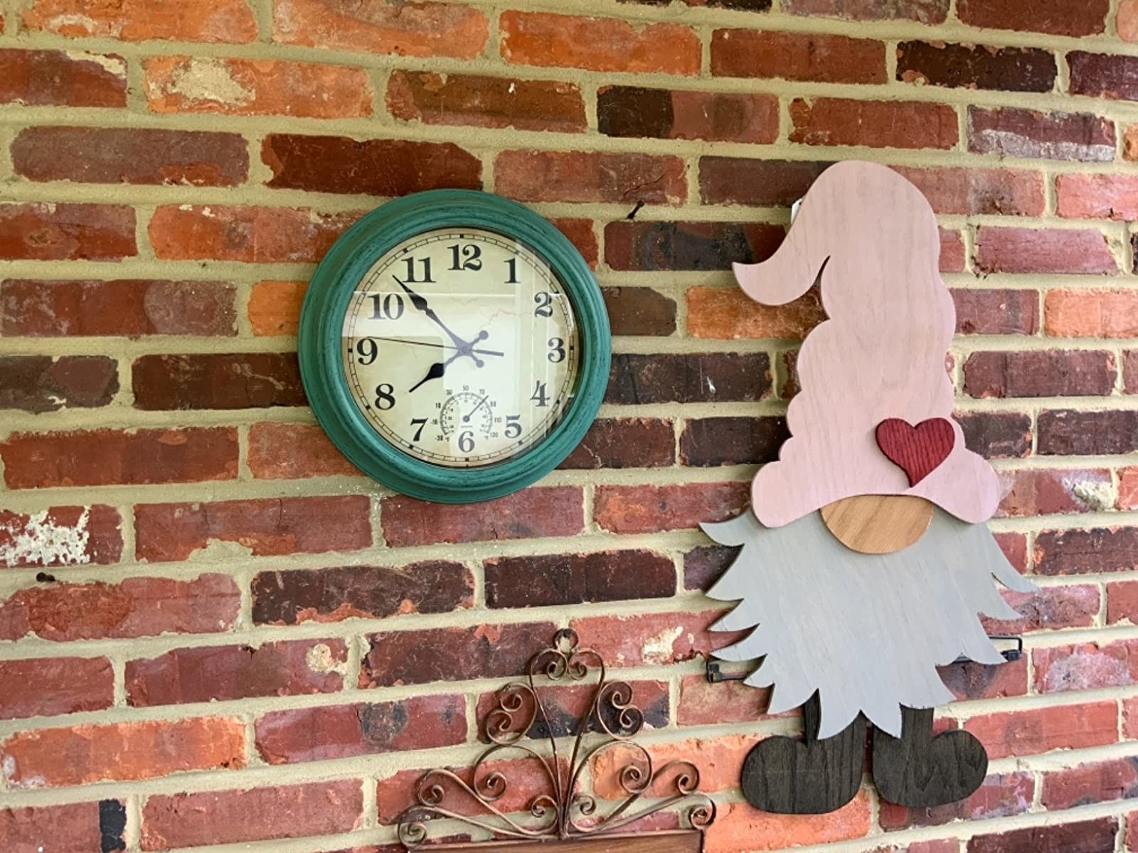 A green outdoor clock next to a gnome wall decor against a brick wall