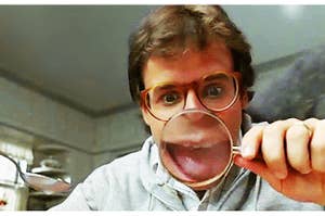 rick moranis has his mouth open behind a magnifying glass so his mouth is magnified and large