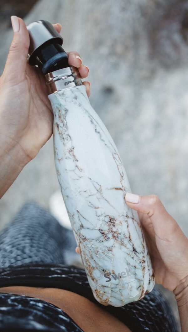 closeup shot of the gold and marble patterned water bottle in a person&#x27;s hand. it has a stainless steel twist-off lid.