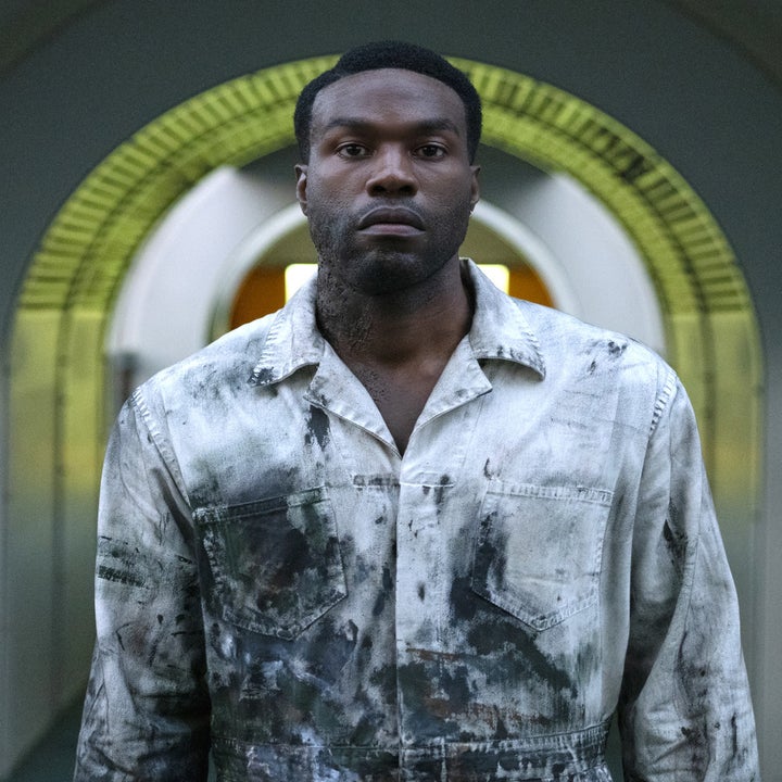 Yahya Abdul-Mateen II in a white painter's jumpsuit covered in black paint, staring at the camera