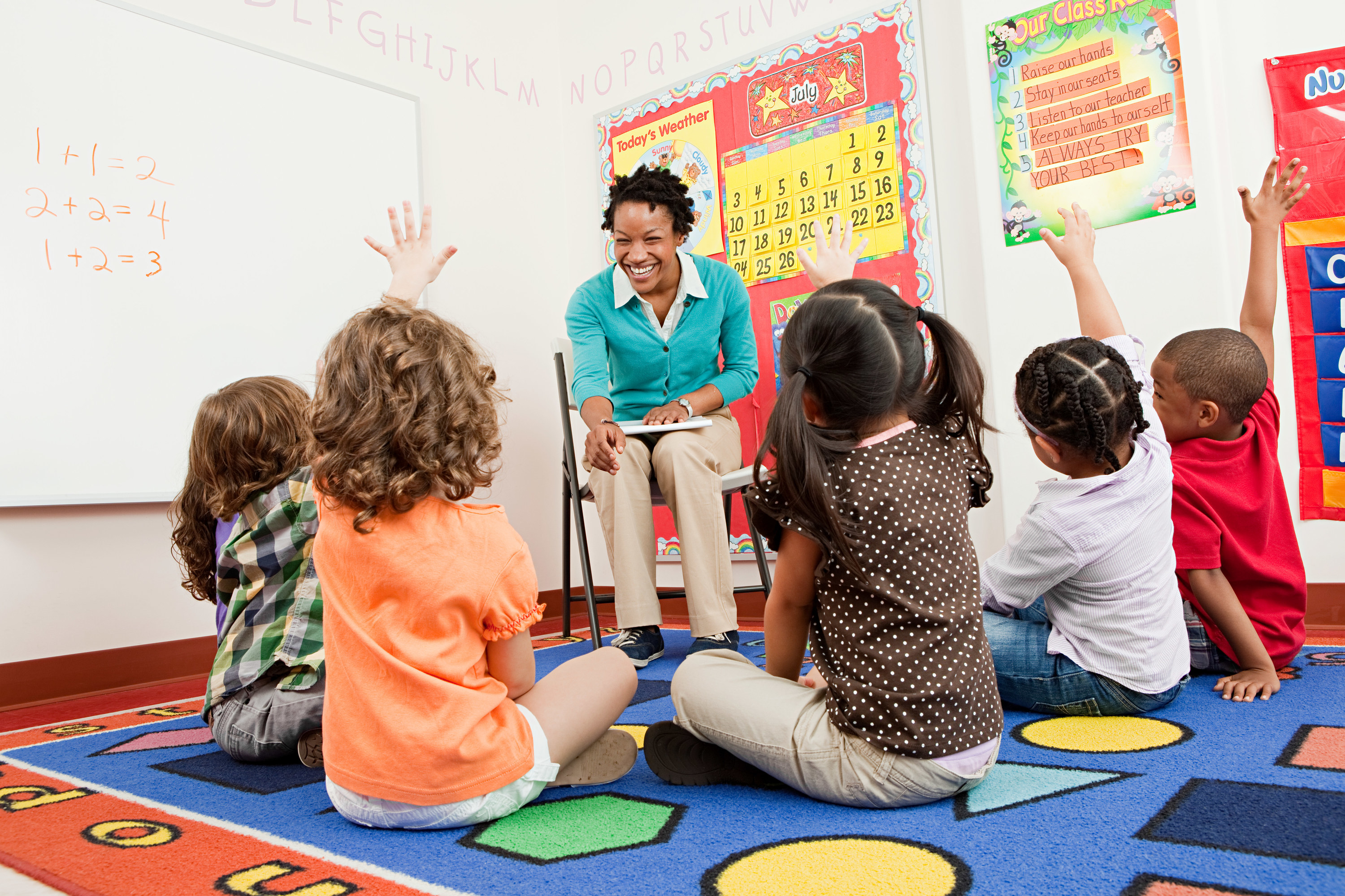 A teacher smiles as her students sit on the rug and raise their hands