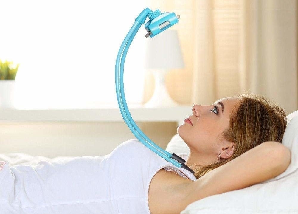A woman lying on a bed with her hands behind her head. She&#x27;s watching her phone which is attached to a neck mount that she&#x27;s wearing