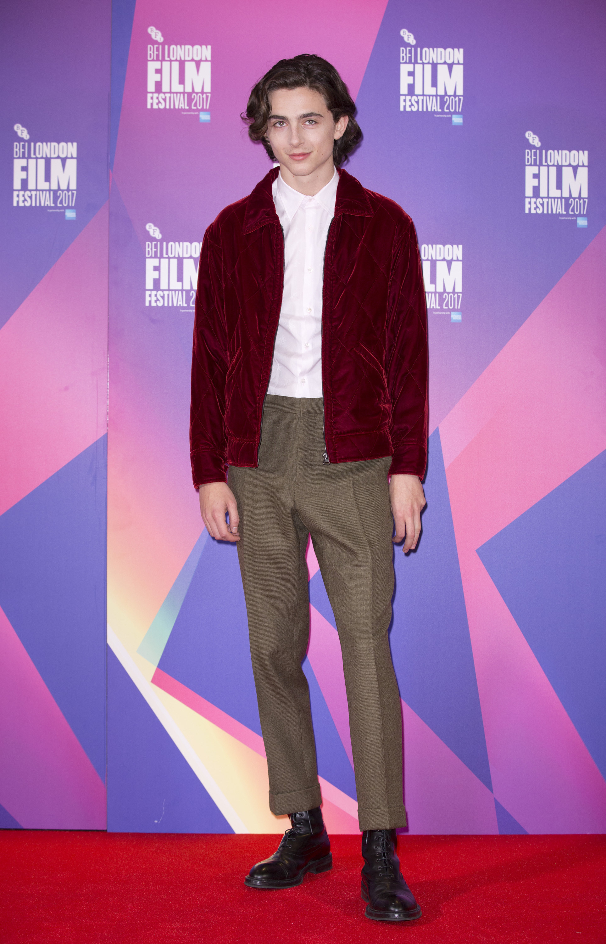 Timothée wears a maroon bomber jacket, white button down, and brown tailored pants