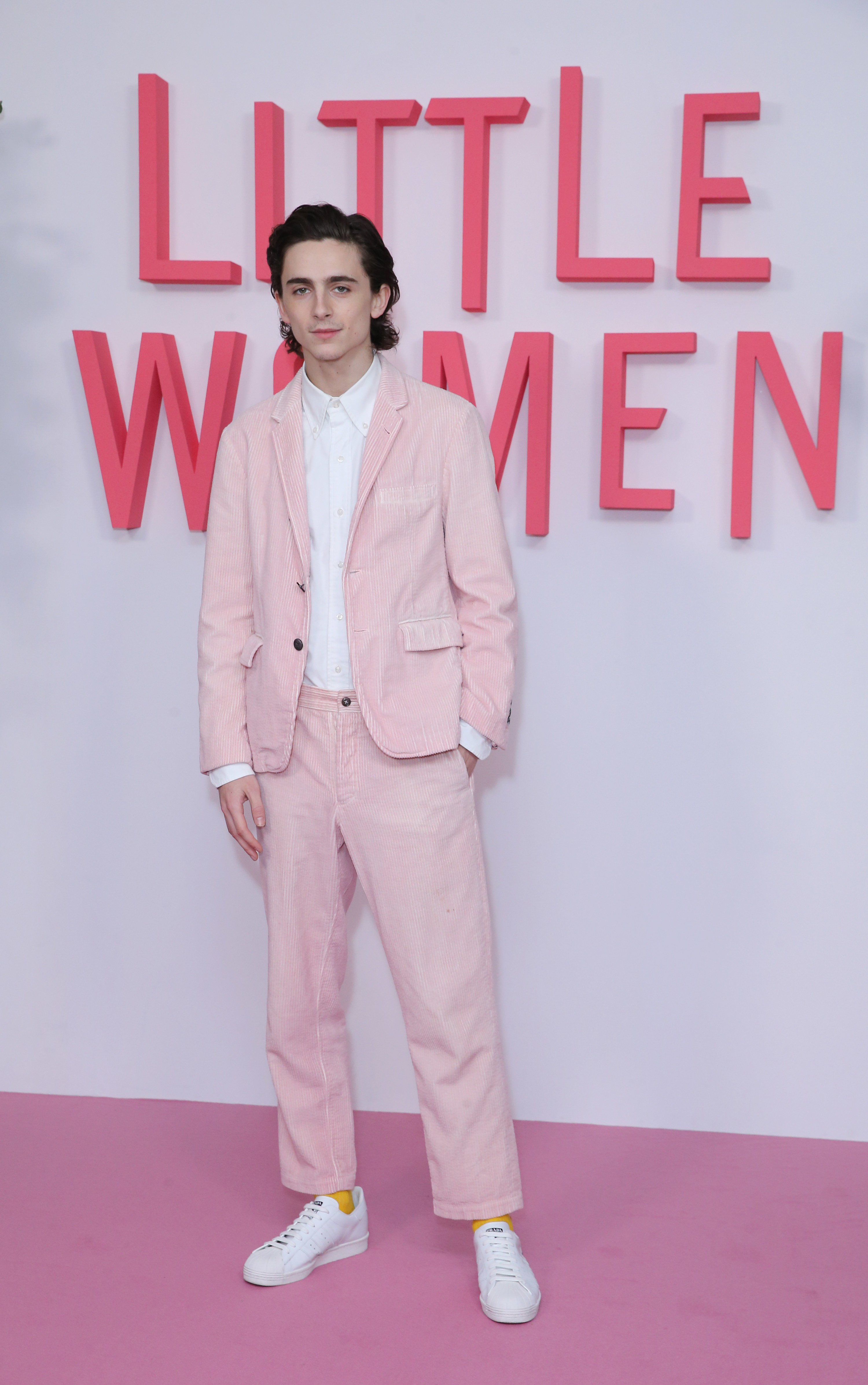 Timothée wears a pink denim suit with white sneakers in front of a light pink backdrop and carpet