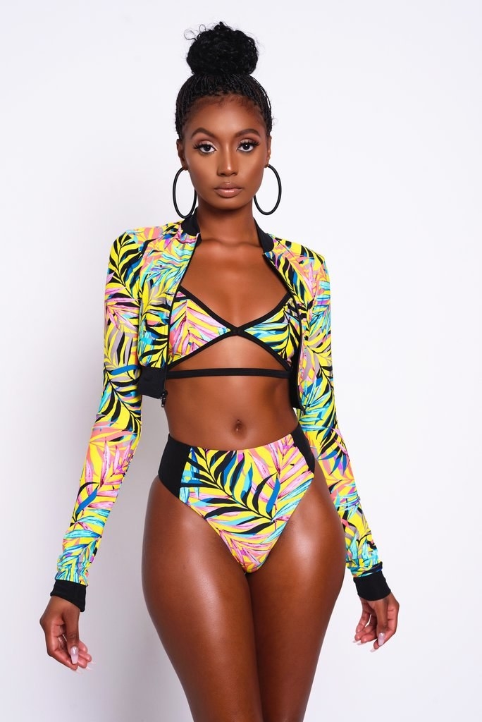 model wearing the bikini top and bottom which have multi-colored palm tree leaves on them and the matching long-sleeve cropped jacket