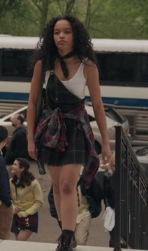 Zoya wears a plaid uniform jumper with one strap buckled to the buttons on the torso and the other hanging loose with a plaid shirt wrapped around her waist