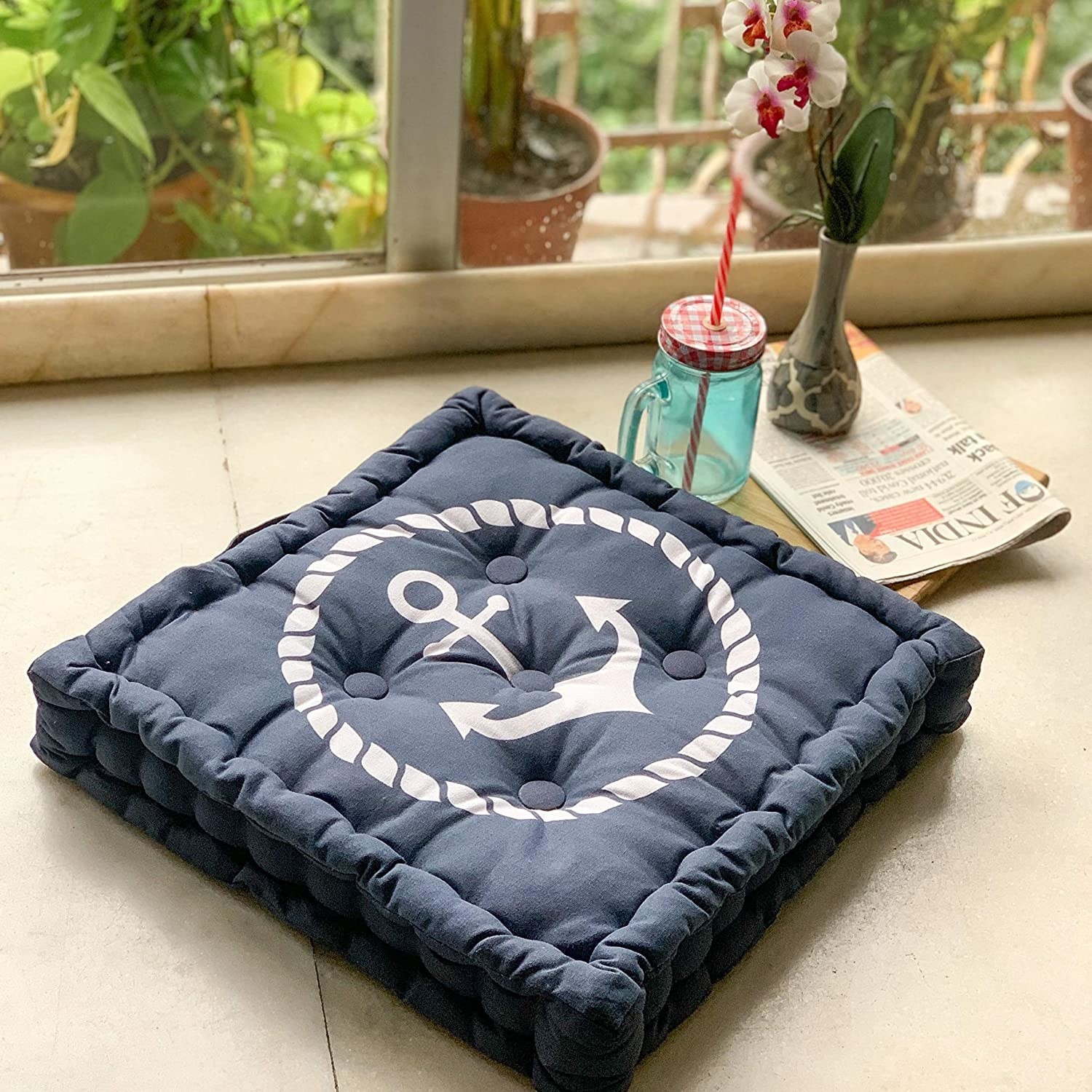 A navy blue square cushion with an anchor printed on it. It&#x27;s kept next to newspapers, a vase of flowers, and a blue mason jar.
