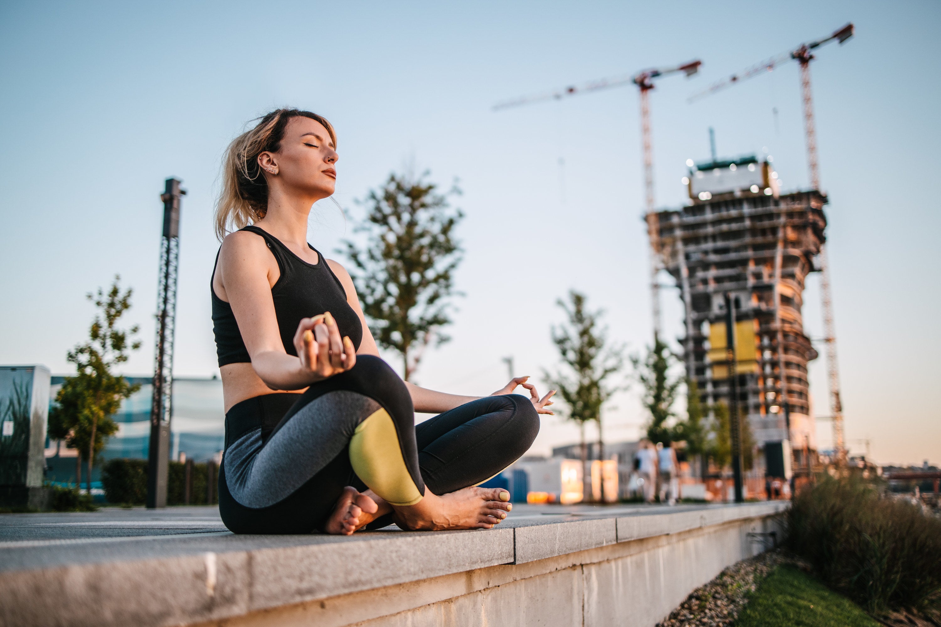 Young adult woman sitting on the ground in the public park meditating next to a construction site