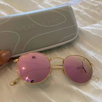 a reviewer photo of a pair of round pink sunglasses next to a hard carrying case 