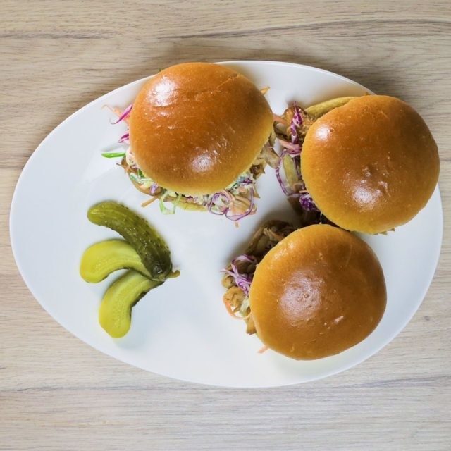An overhead shot of three pork sliders, with the slaw oozing out from the brioche bun edges, and a garnish of three halved gherkins.