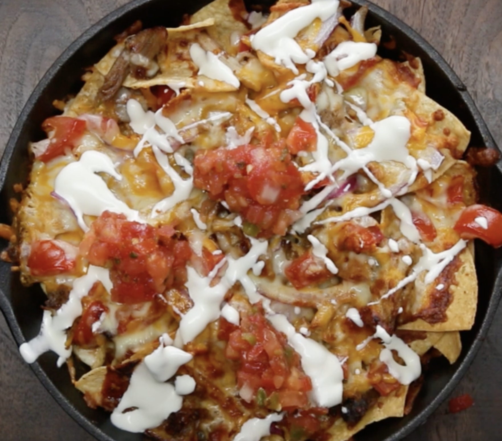 Overhead shot of a cast iron pan of baked nachos with pulled pork, tomato salsa and a sour cream drizzle.