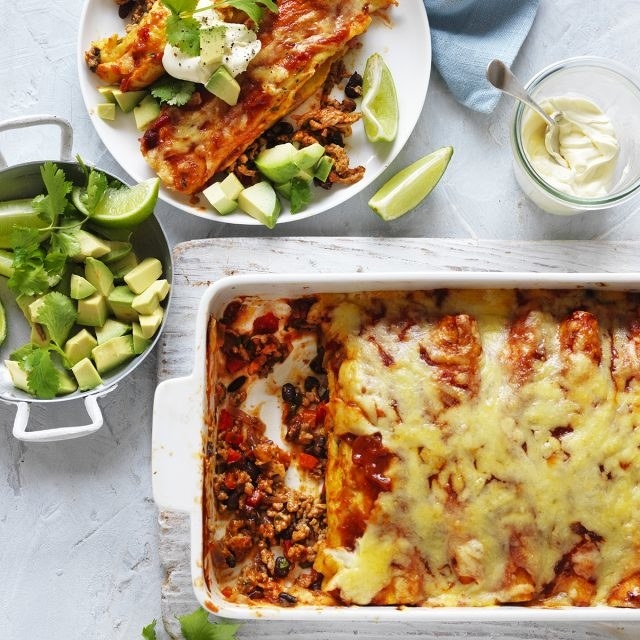 Oven dish of enchiladas with one portion already plated and a side of diced avocado.