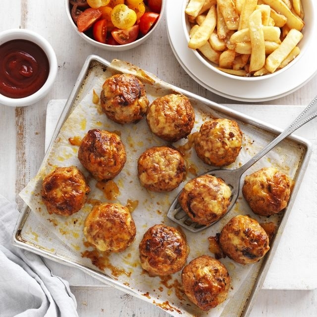 Twelve pork rissoles on a baking sheet on an oven pan, with sides of sauce, halved cherry tomatoes and a small bowl of fries.