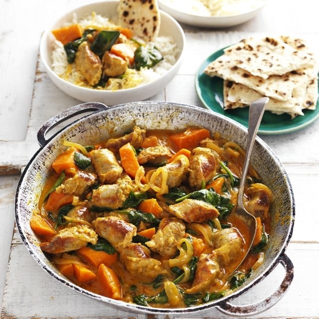 A korma dish with sausages, sweet potato and spinach with a small plate of naan at the back, and a bowl of rice topped with korma.