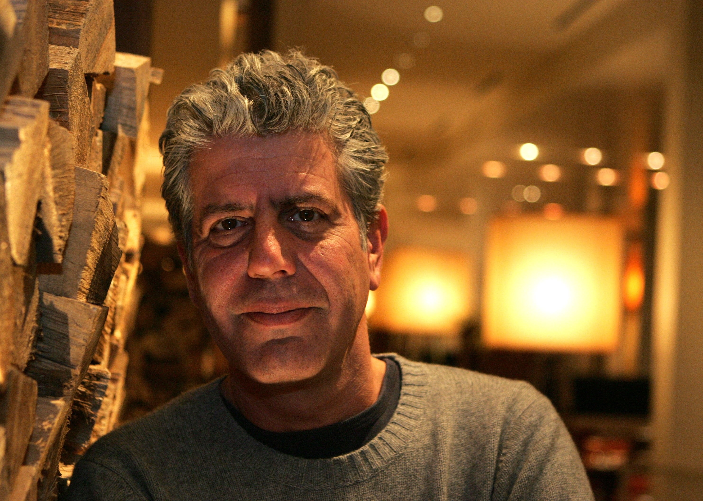 Closeup photo of Anthony Bourdain in a restaurant leaning against a woodpile and looking at the camera