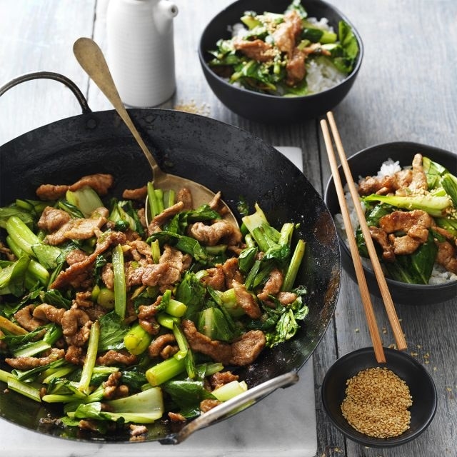 A wok with a greens and pork stir fry and two bowls of rice topped with the stir fry.