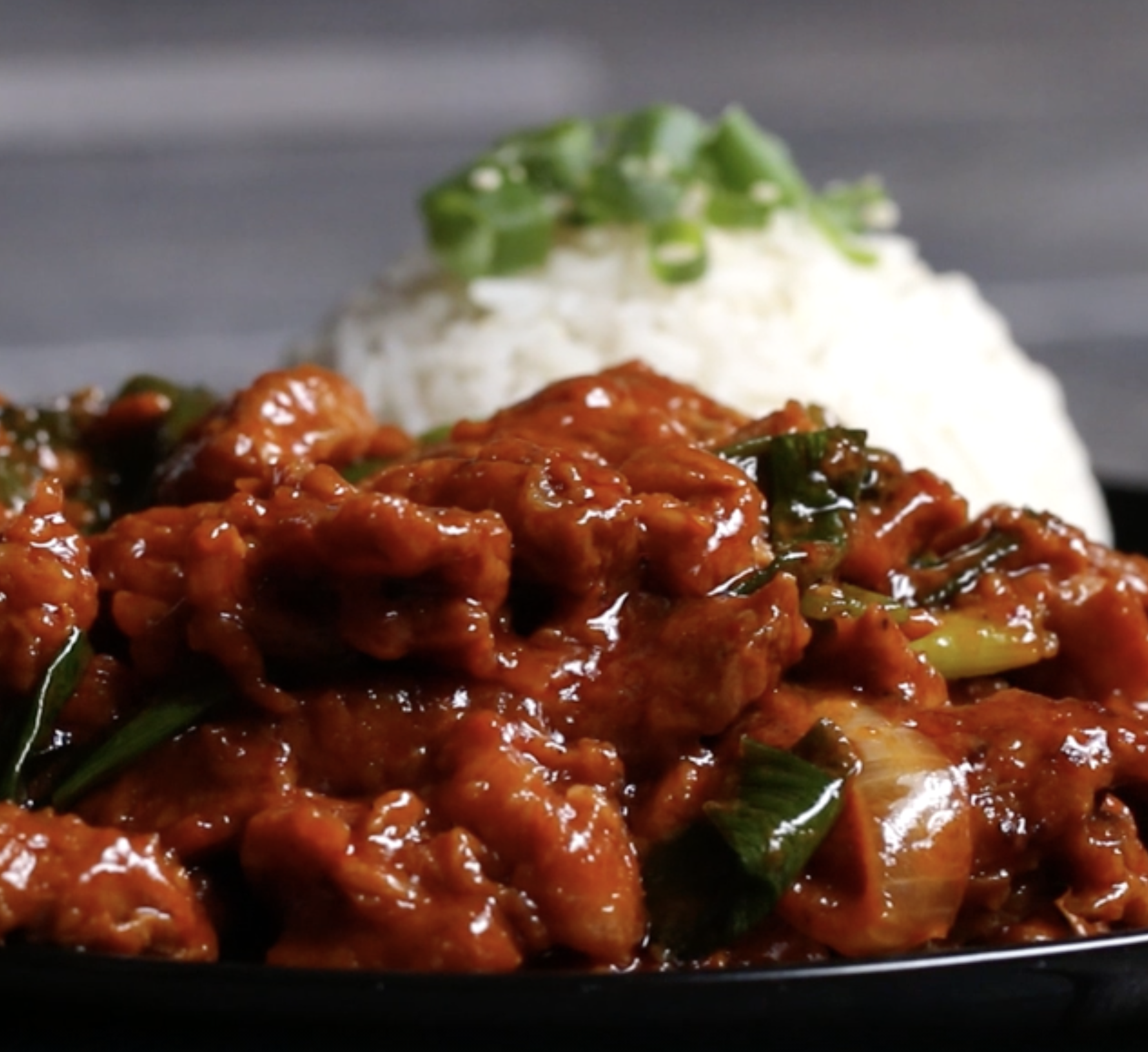 Close up of spicy Korean-style pork with a bed of rice topped with spring onions in the background.