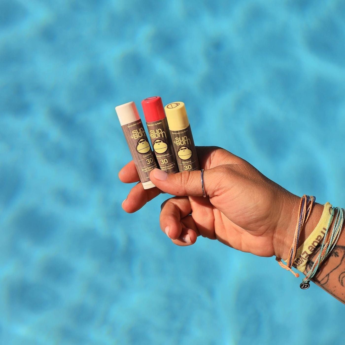 A hand holding three lip balms over a pool