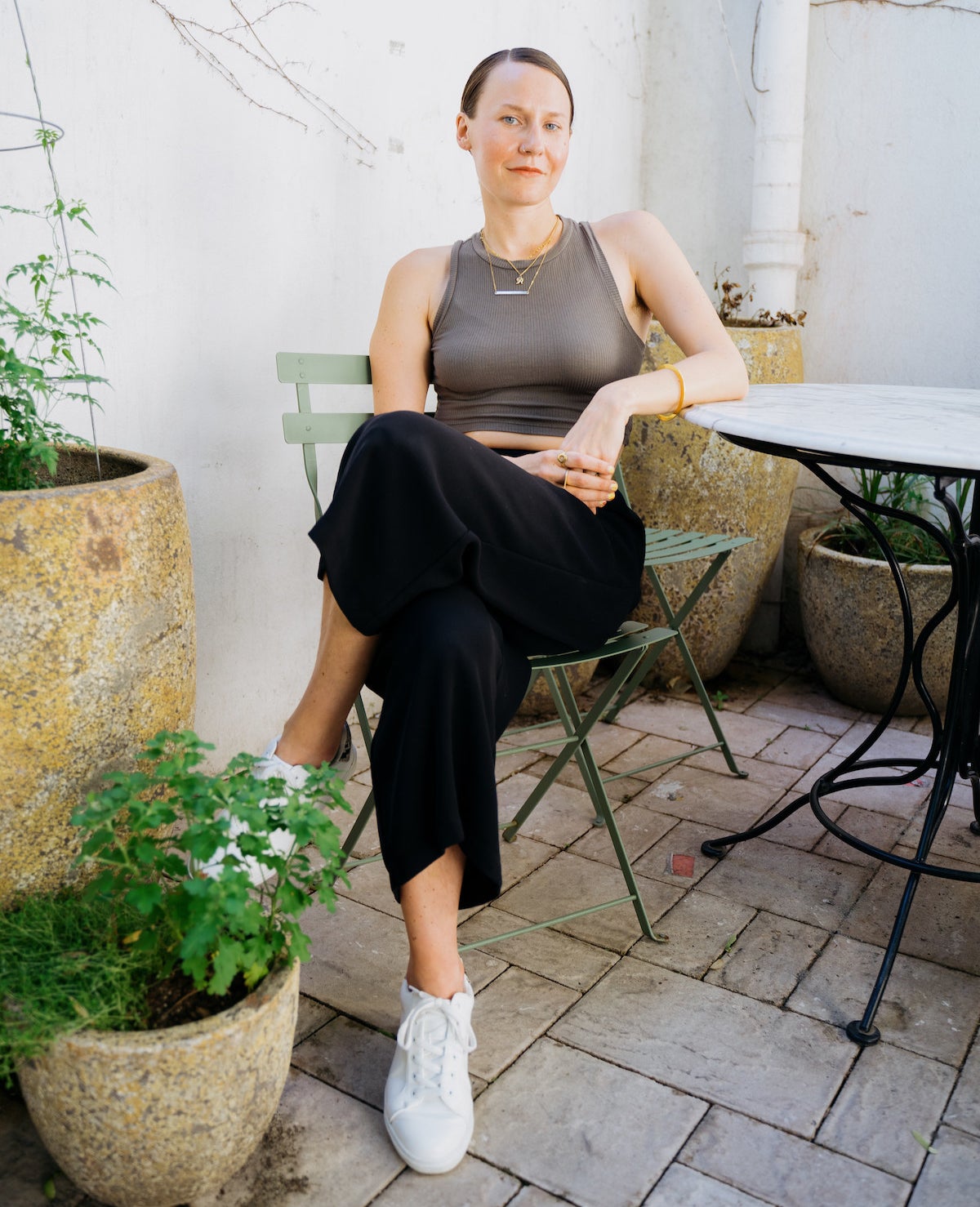 A woman wearing pants and sneakers sits at a table with her legs crossed and her elbow leaning on the table