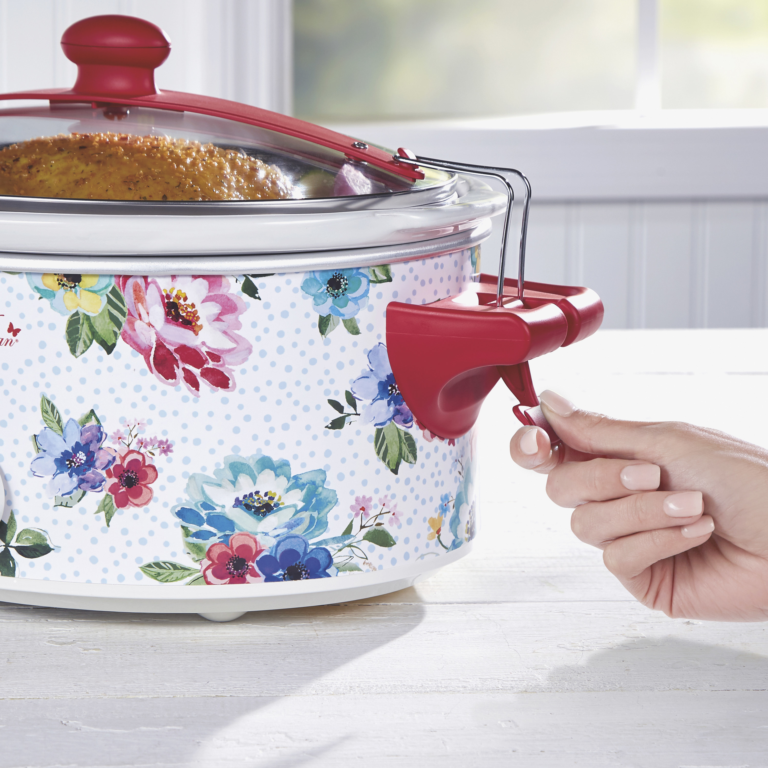 A model demonstrates the clip on the slow cooker, which ahs red and blue flowers on a white background on the side and red clips and handles.