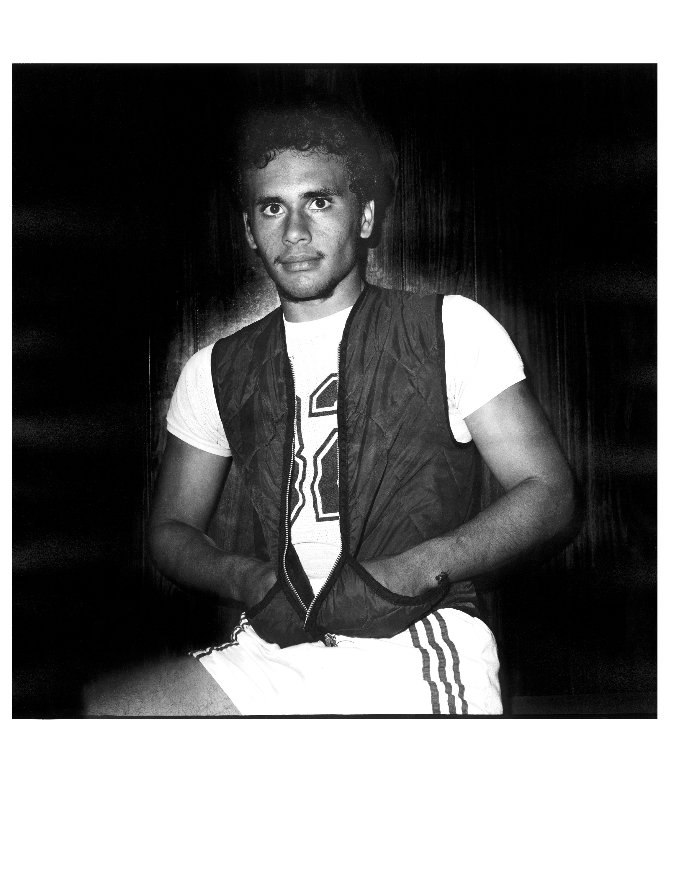 A young man in basketball shorts and a vest looking straight at the camera