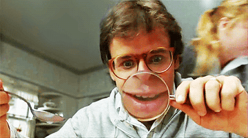 rick moranis talks, facing a magnifying glass, so his mouth is large and magnified
