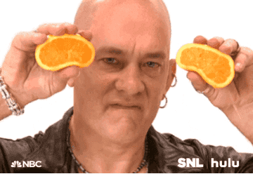 Bald Tom Hanks with hoop earrings looking into the camera and squeezing juice out of orange halves