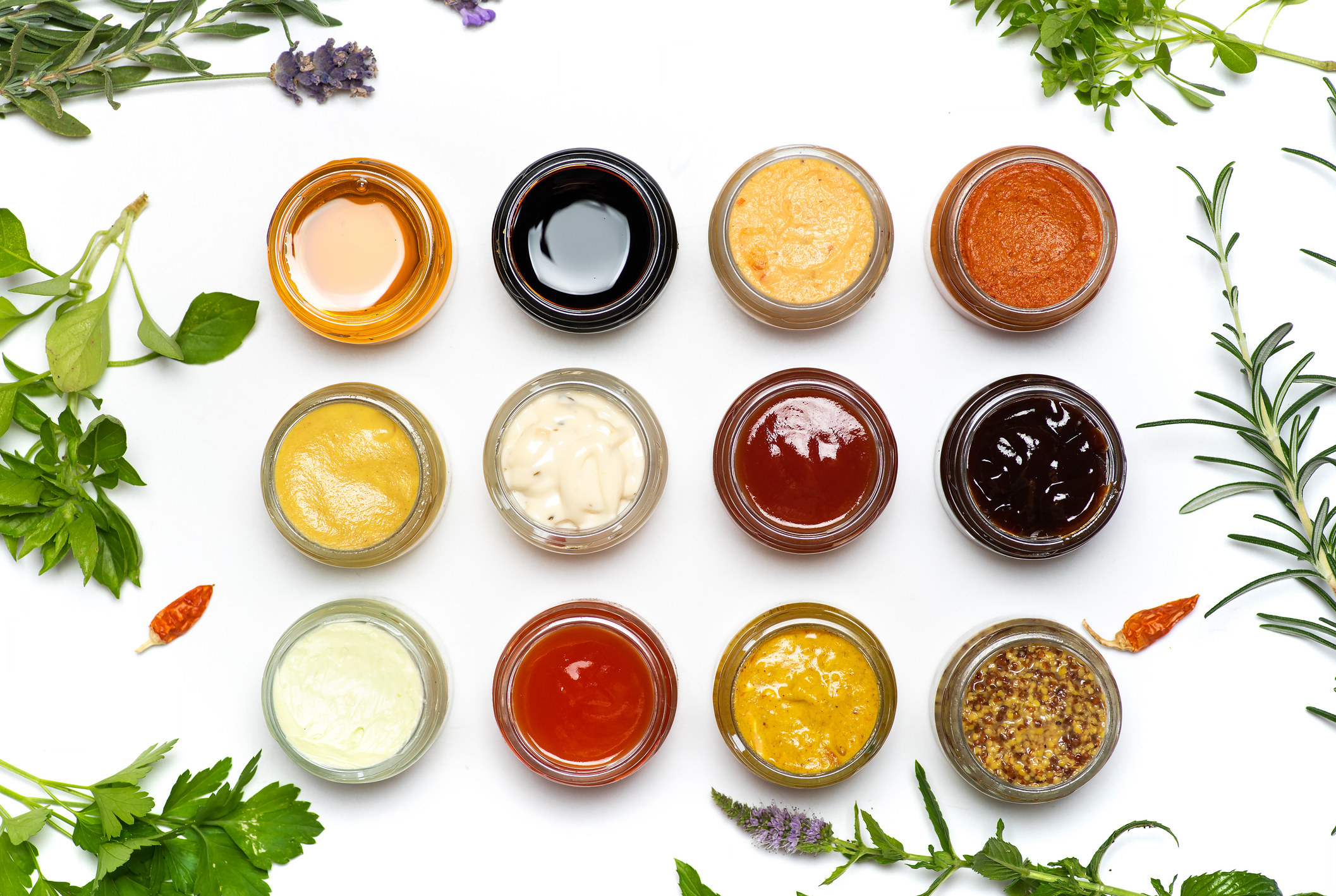 Twelve ramekins of various sauces arranged in rows with fresh herbs at the sides of the frame