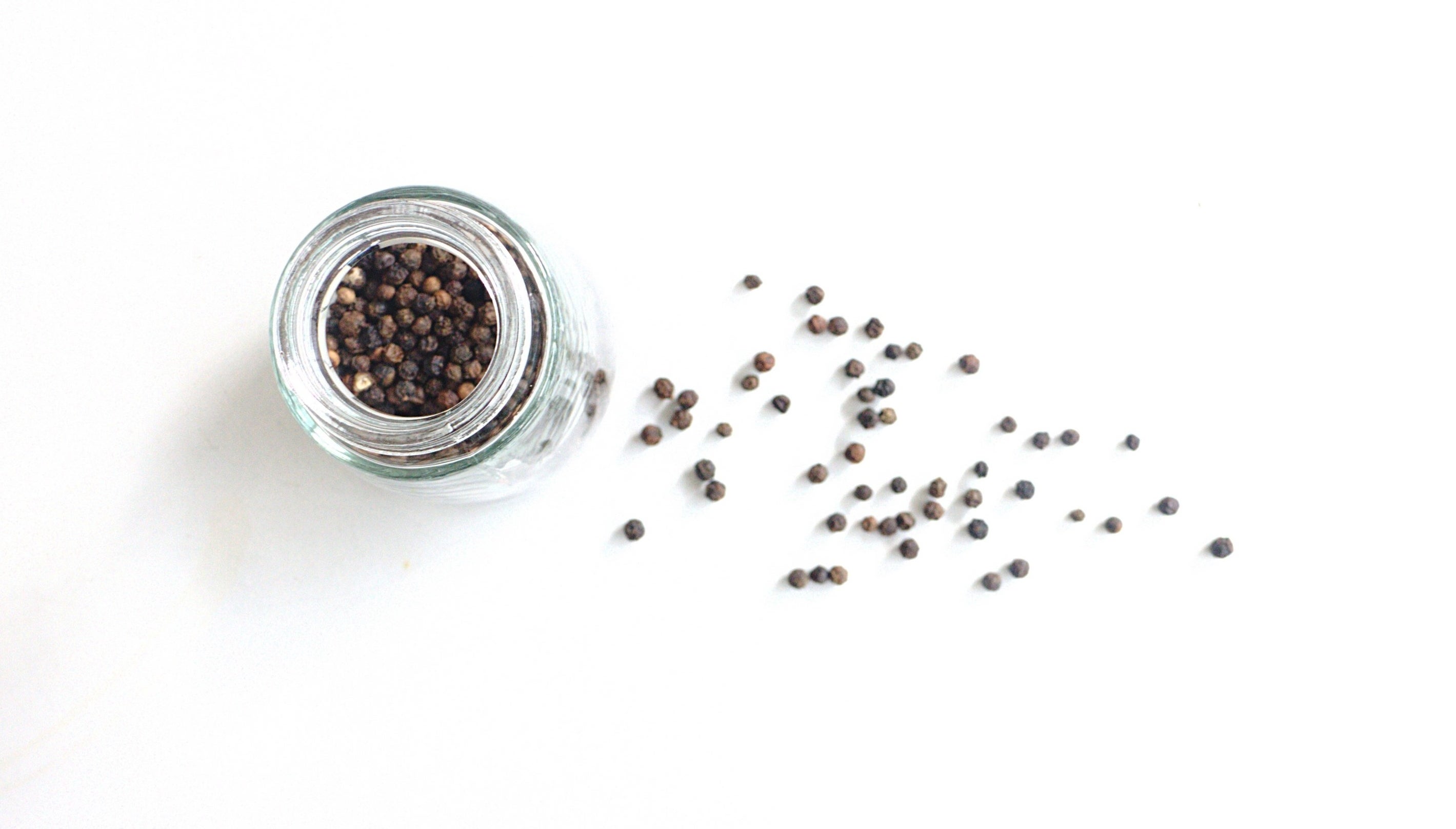A top view of peppercorns in a small, unlidded glass container with several peppercorns strewn to the side