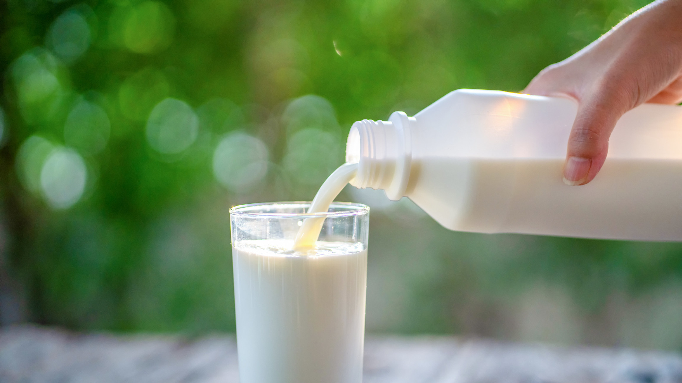Milk being poured out of a narrow plastic jug into a glass; greenery in the background