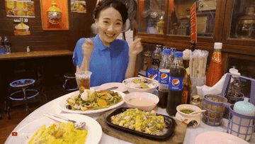 A lady smiling, holding a fork in each hand, sitting in front of a table with several dishes, tons of condiments, and chopsticks