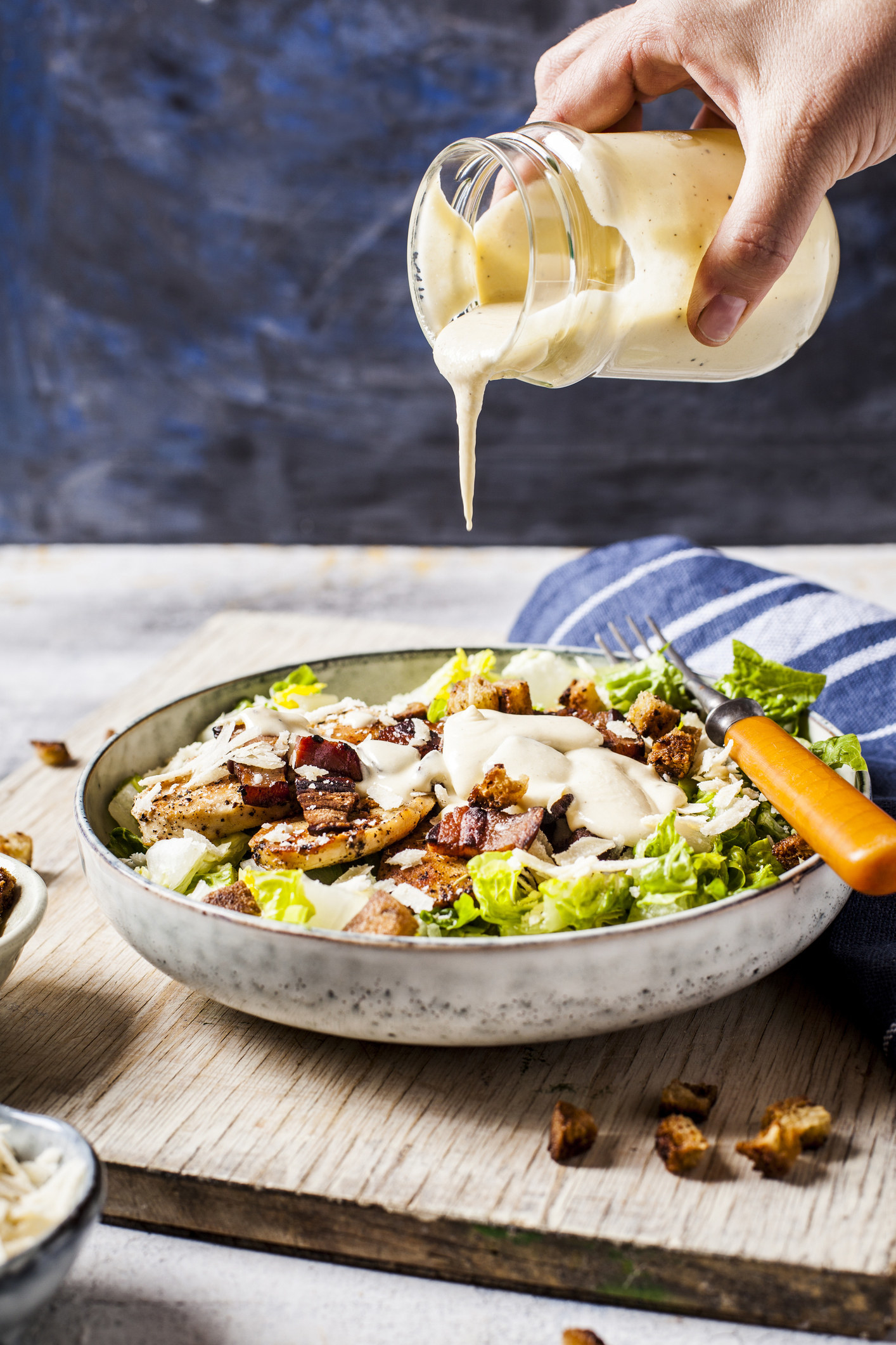 A hand pouring a thick Caesar dressing out of a glass jar onto a fresh salad in a bowl on a wooden board