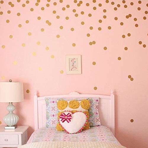 A pink wall with golden polka dot decals. There&#x27;s a bed with cushions and a side table with a lamp on it.