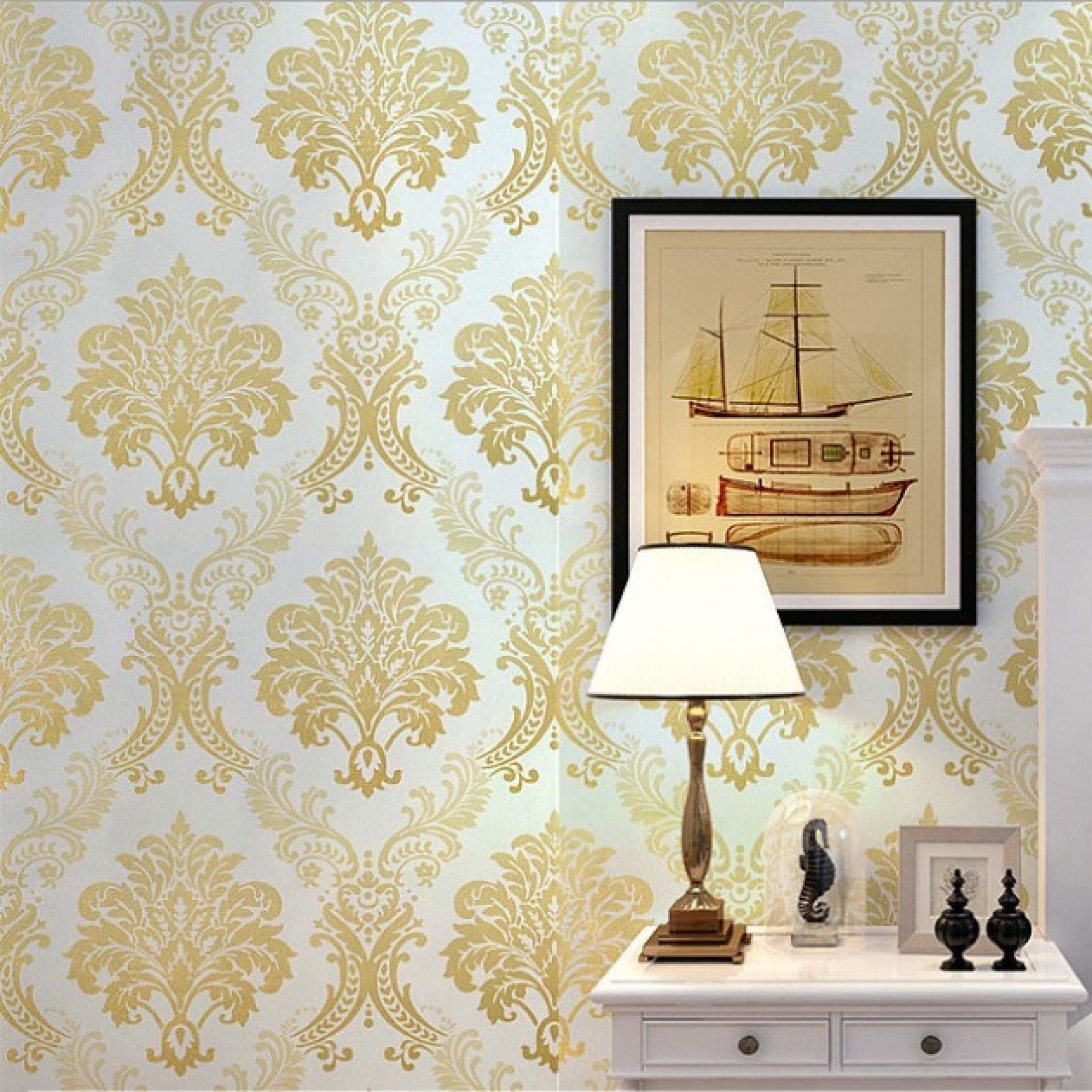 A white and golden floral wallpaper on a wall. There&#x27;s a white table placed in front of it, with a lamp and other showpieces. There&#x27;s a framed painting of a boat on the wall.