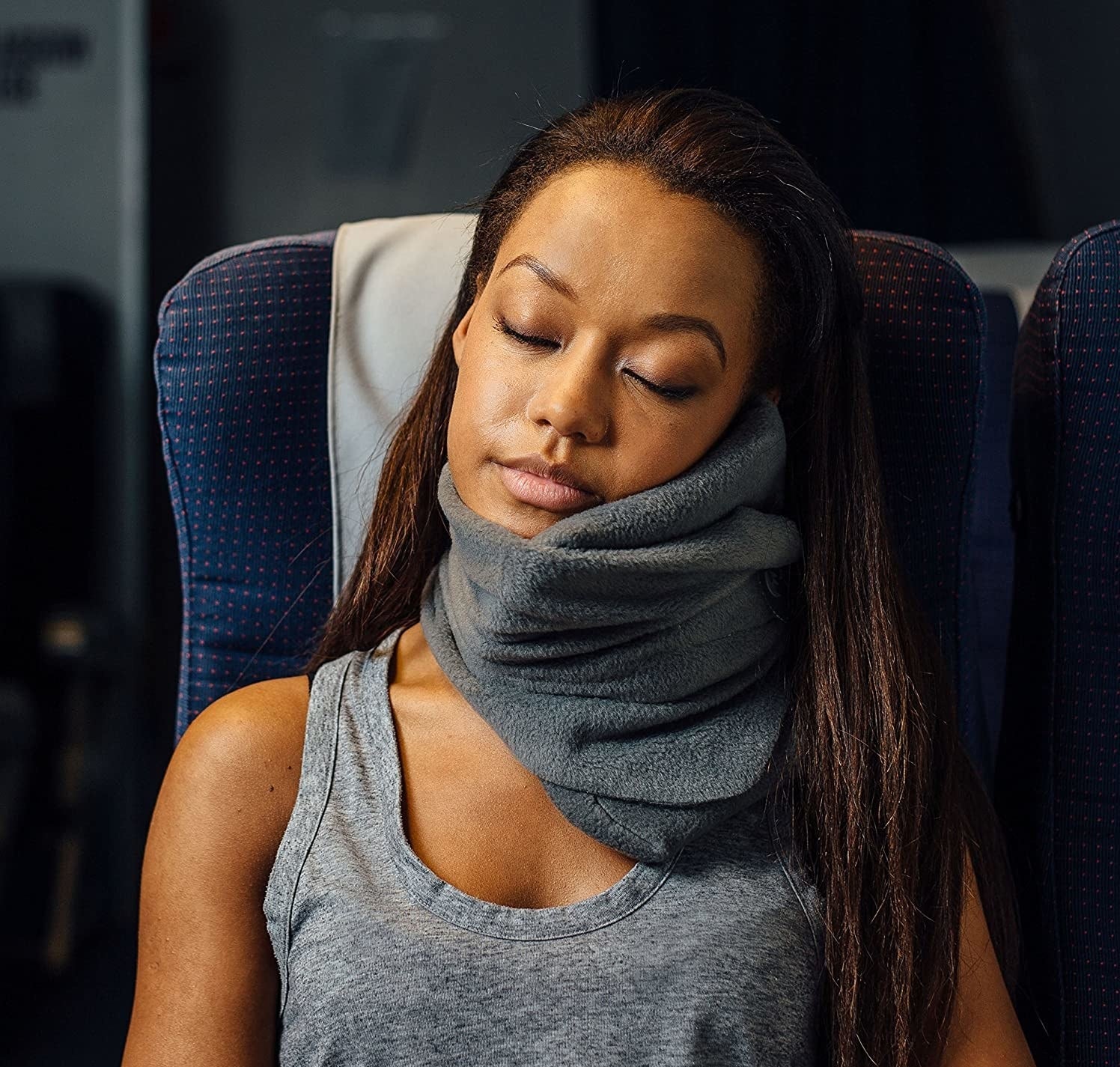 A model wearing a gray Trtl pillow while sleeping on a plane