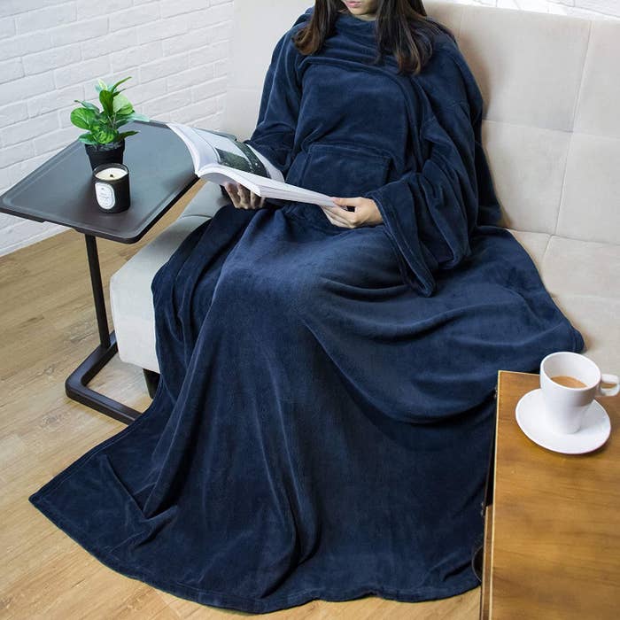 A woman sitting on a living room couch reading a magazine while wearing a sleeved fleece blanket