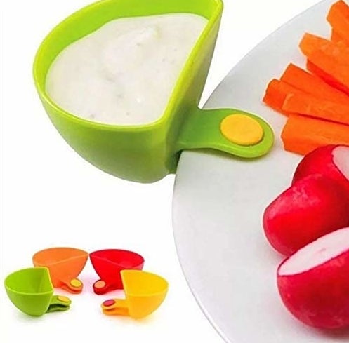 A filled dip clip attached to a plate that&#x27;s carrying fruits and vegetables, next to four other dip clips in multiple colours