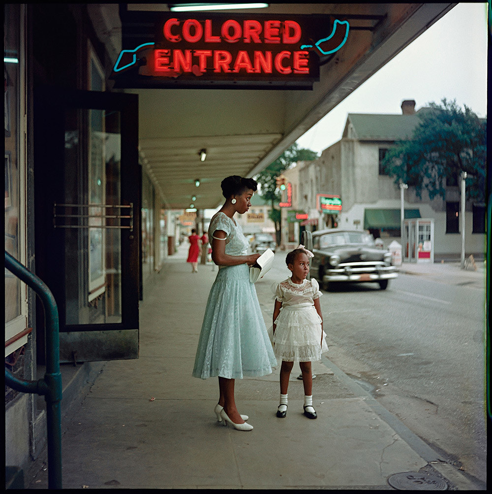 A mother and child in dresses stand under a &quot;colored entrance&quot; sign