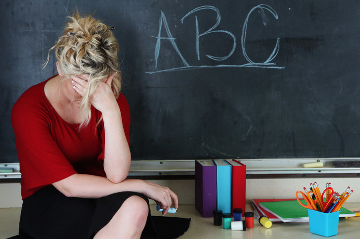 Elementary classroom setting with tired or frustrated teacher holding her head. She&#x27;s sitting in front of an chalkboard with ABC