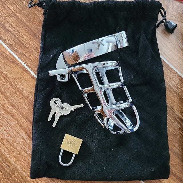 Silvertone chastity cage, three keys and goldtone lock on top of black storage bag