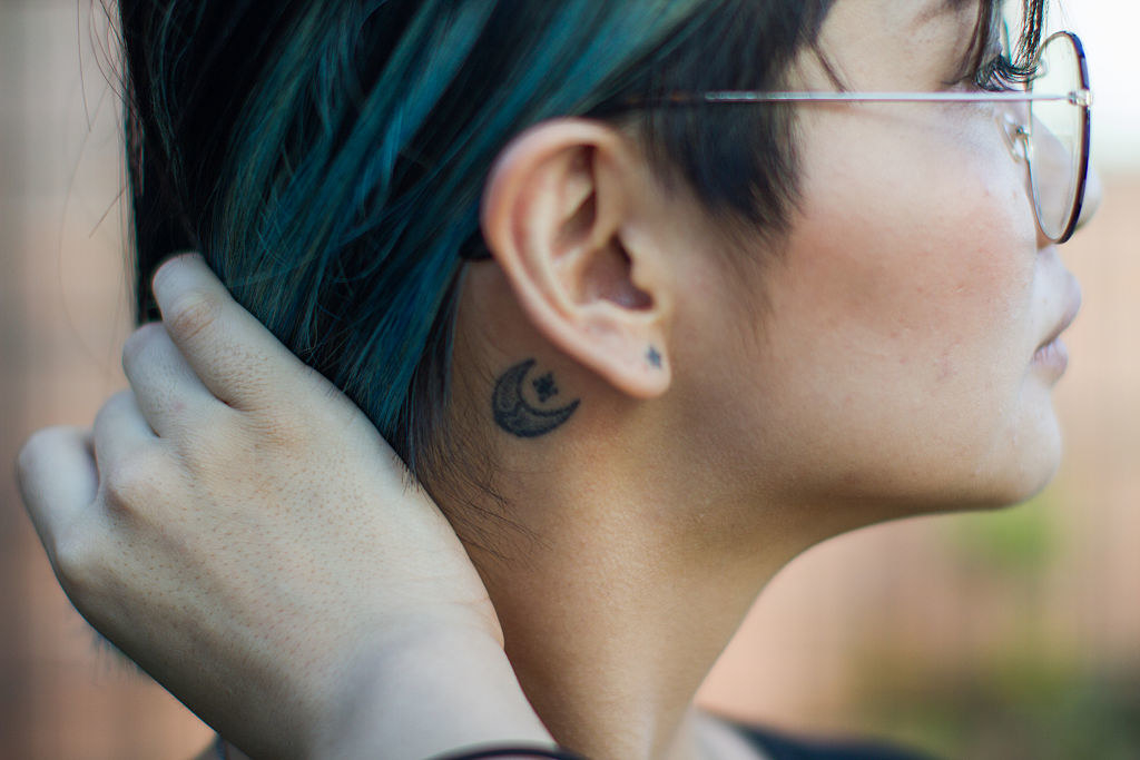 A woman pulls back her hair to reveal a tattoo of a crescent moon and star behind her ear