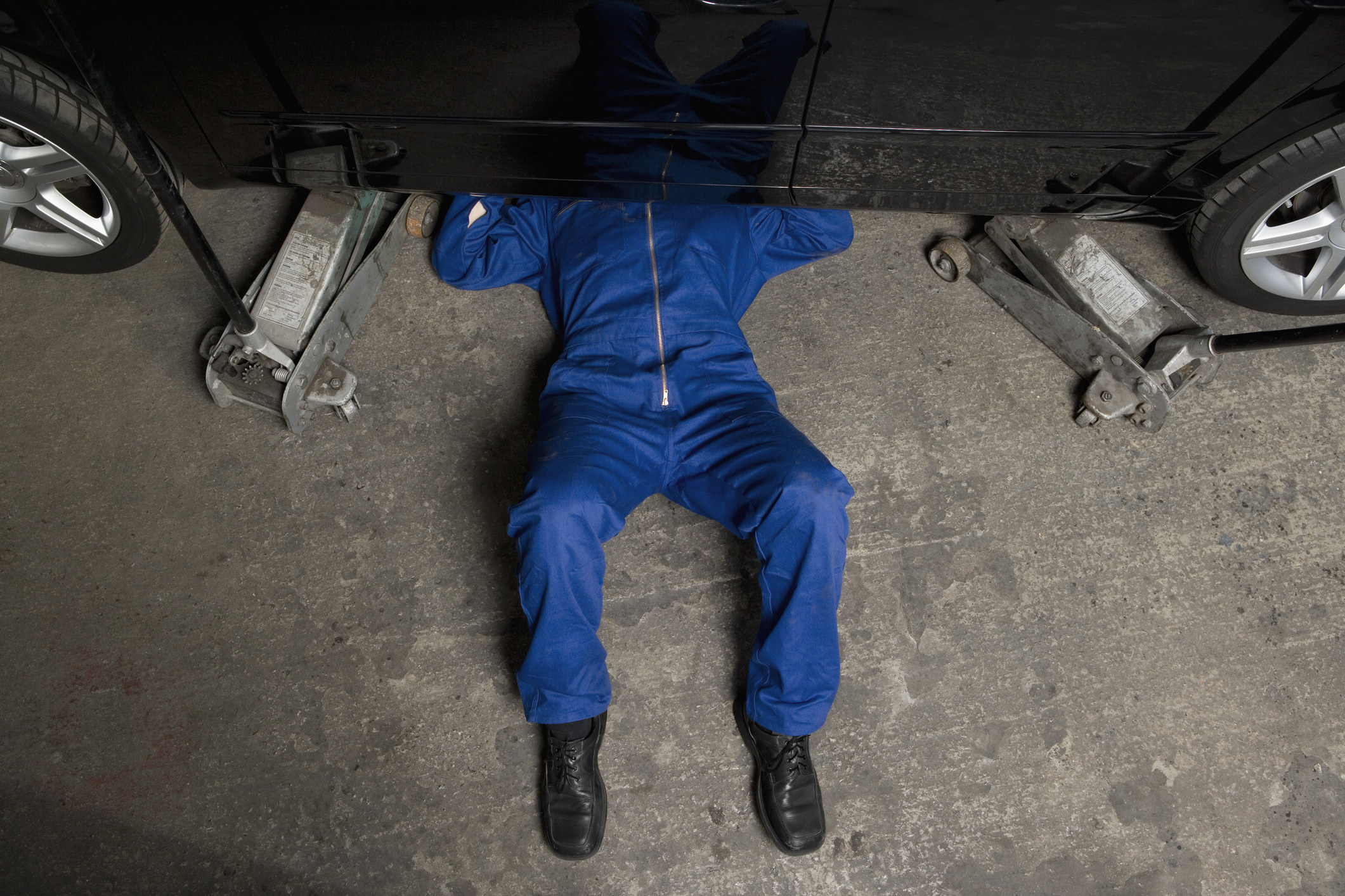 Top view of the torso and legs of a person in a worksuit and boots underneath a vehicle supported by jacks on either end