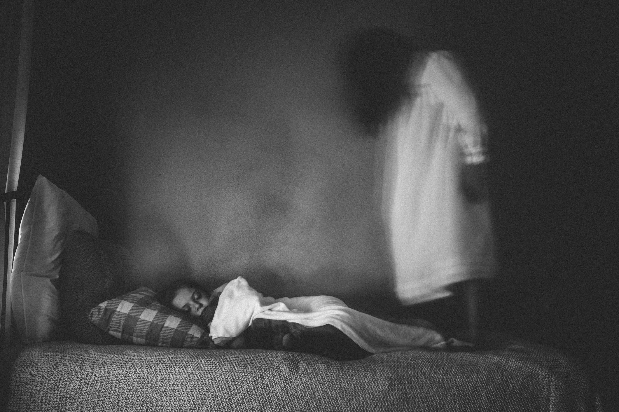 A girl sleeping in bed with a ghostly figure of a woman in a nightgown and dark hair covering her face, standing on the end of the bed above the girl&#x27;s feet