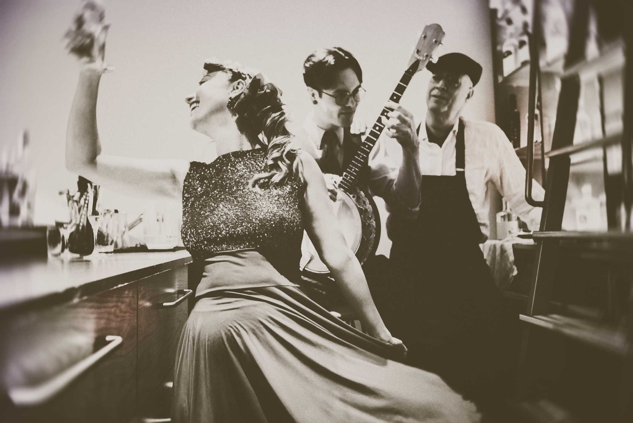 A sepia photo of a woman in party dress holding up a glass to cheers, behind her a man with shiny, parted hair and glasses plays a banjo, and just behind him stands another man in overalls and a cap