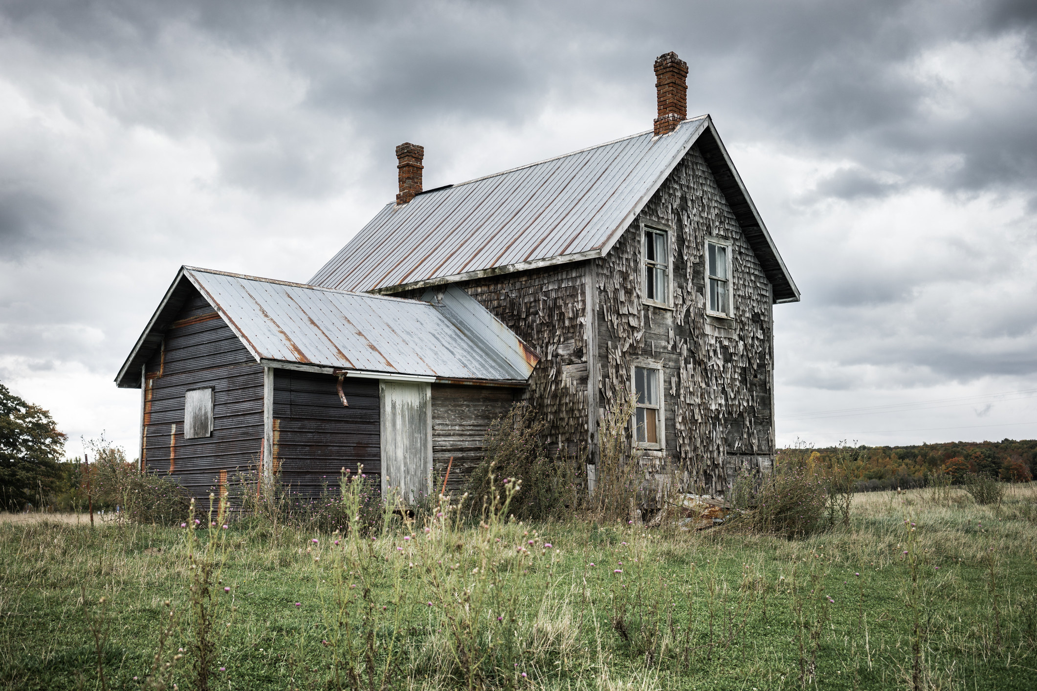 An old house with wooden siding and two chimneys in a field with ominous clouds behind