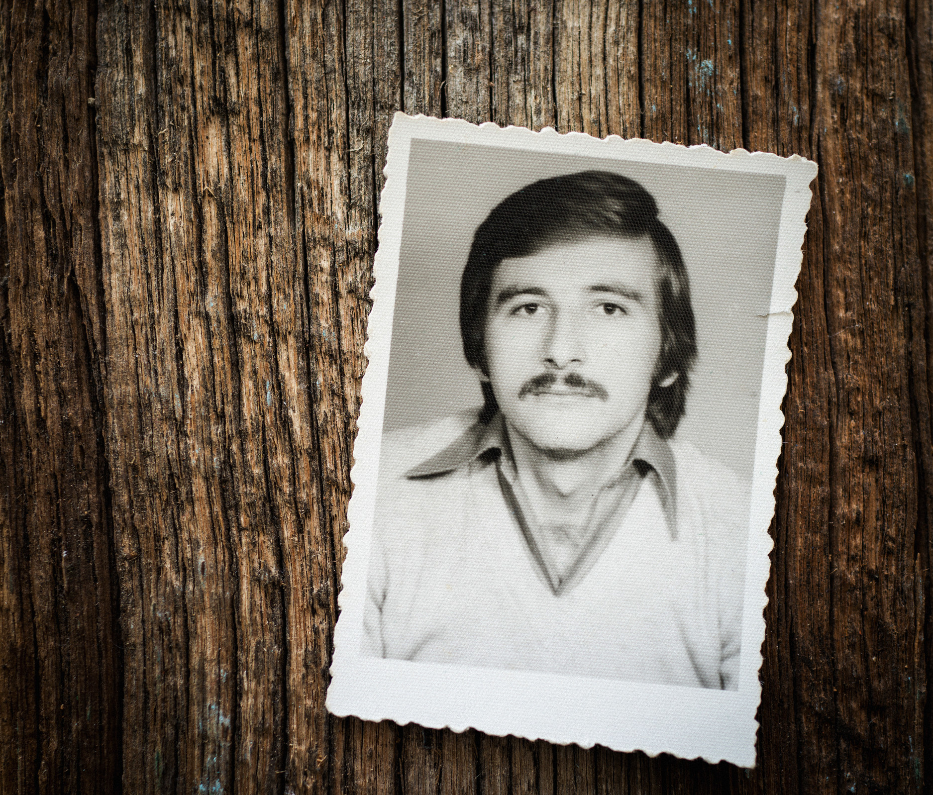A vintage photo on a piece of wood, showing a man with side-parted hair and mustache wearing a large-collared shirt underneath a v-neck sweater