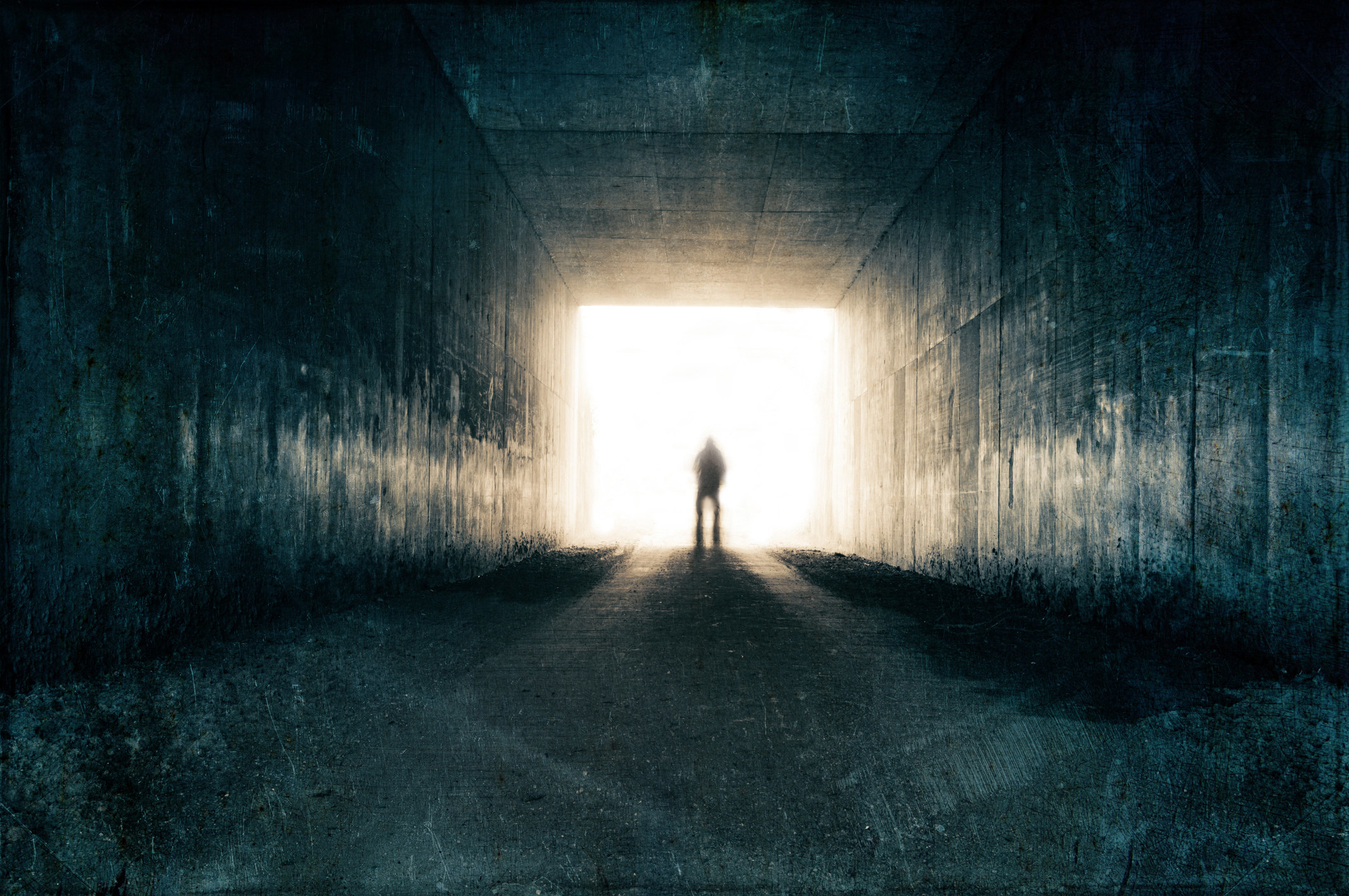 A dark walkway with a bright light at one end, within it is the dark silhouette of a man