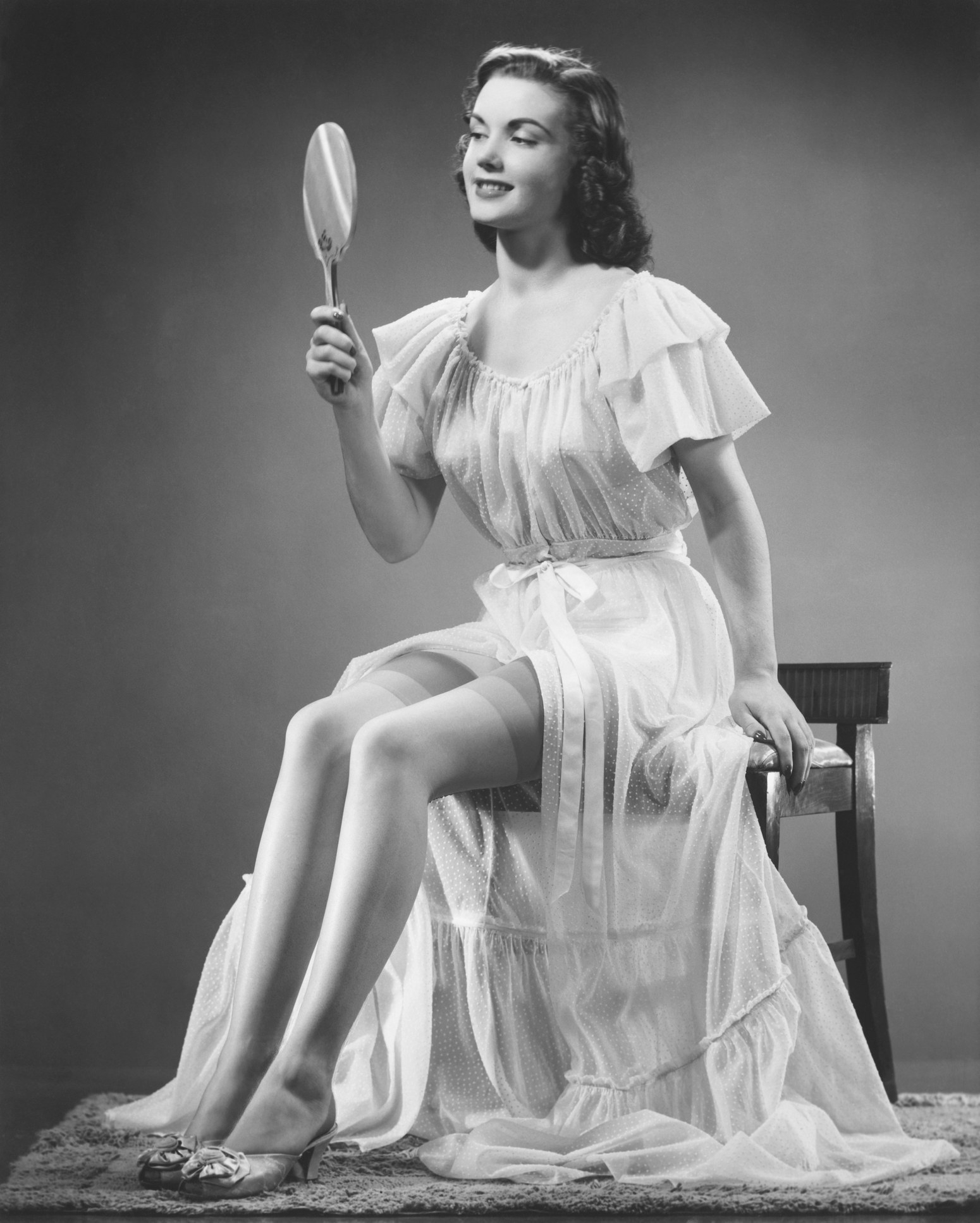 A &#x27;30s styled woman perched on a lowback chair wearing a long, flowing nightgown with short sleeves and high-heel slippers, looking into a hand mirror